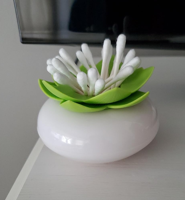 A white container with a modern design, holding cotton swabs arranged to form a flower, placed on a white surface beside a black object.