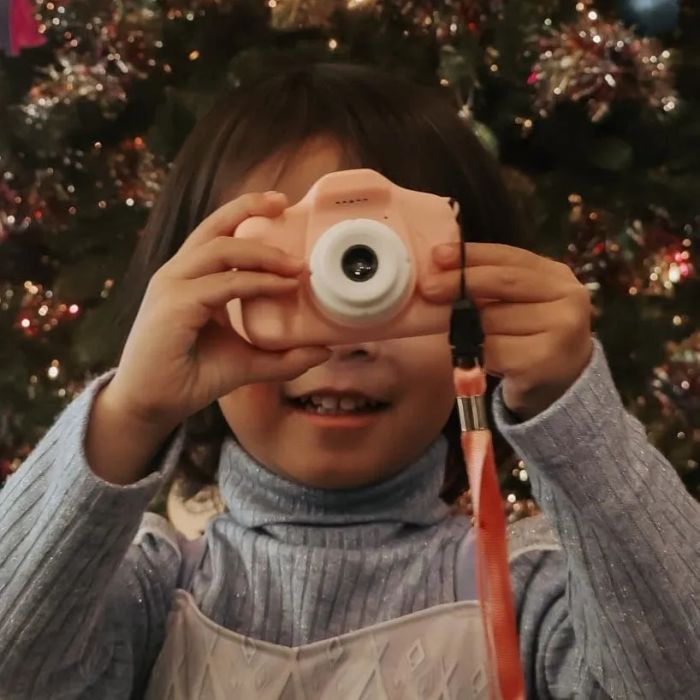 A child standing in front of a Christmas tree, holding a pink camera and wearing a dress featuring Elsa from Frozen