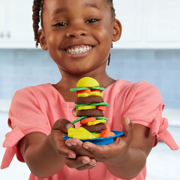 A child grinning as they proudly hold up a colorful burger stack made from modeling clay, set in a playful shopping environment