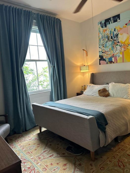 A modern bedroom featuring a spacious window with blue curtains, abstract wall art, a bed dressed in white linens with a blue throw blanket, and a pendant lamp hanging above the bedside table, creating a stylish and serene atmosphere.