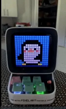 A compact speaker featuring a screen displaying a dancing penguin pixel graphic, with a mini keyboard and six colorful buttons