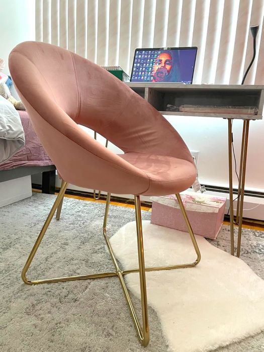 A pink modern accent chair with gold legs, placed in a room featuring a desk, laptop, and decorative items, showcasing a chic home office environment.