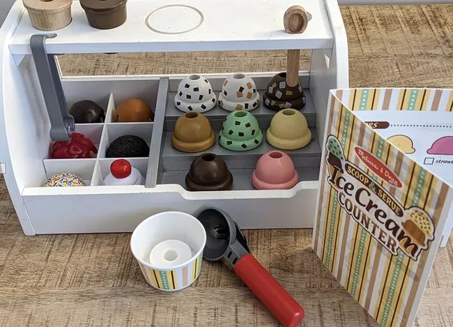 A wooden ice cream playset complete with a variety of scoops, toppings, and a menu displayed on a charming wooden counter