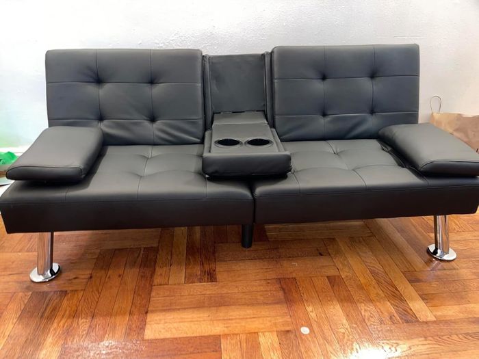 A stylish black leather futon with integrated cup holders, armrests, and chrome legs, set on a wooden floor.