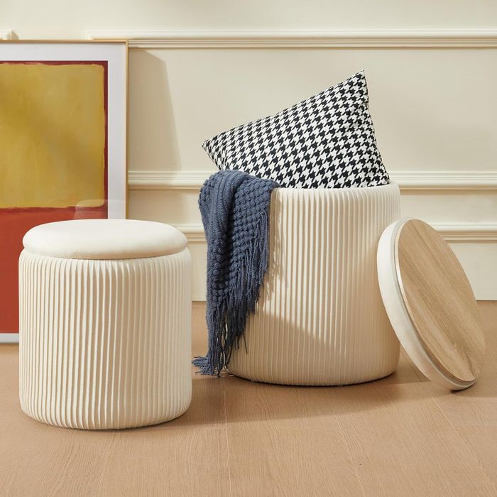 Cream-colored ribbed storage ottomans, one containing a houndstooth pillow and a blue blanket, set on a light brown wooden floor.