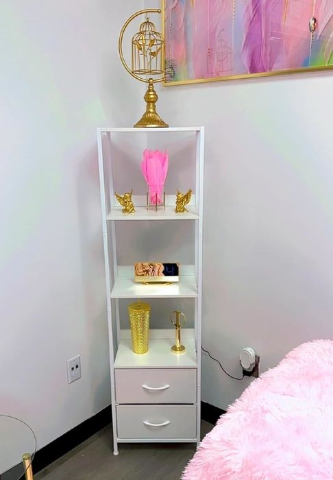 A white shelving unit adorned with decor items such as a gold lamp, small sculptures, books, a vase, and a clock, positioned next to a pink-covered bed and a glass side table.