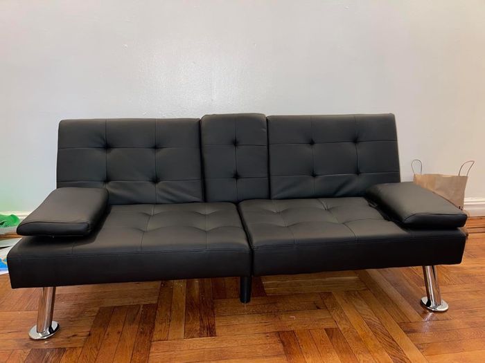 A sleek, contemporary black futon sofa featuring stainless steel legs, positioned on a wooden floor with a paper shopping bag placed nearby in the background.