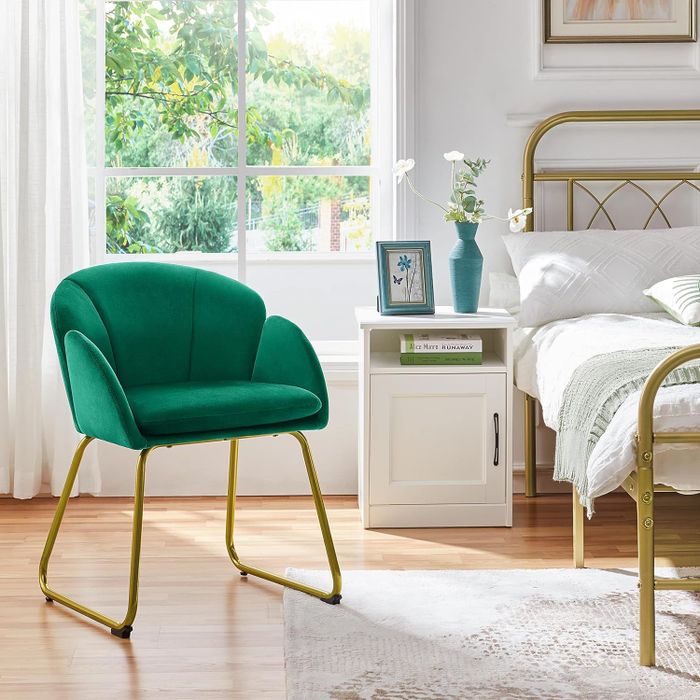 A green accent chair with gold legs positioned in a bedroom beside a white nightstand.