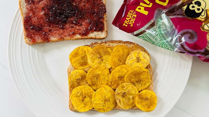 A picture of a deconstructed peanut butter and jelly sandwich with plantain chips on a plate. One bread slice is spread with jelly, while the other has peanut butter topped with plantain chips.