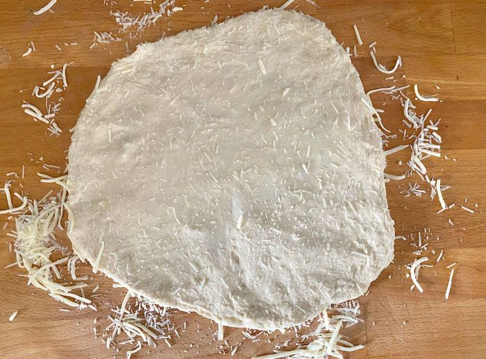 On the counter, there's shredded cheese, with pizza dough placed on top, its edges stretched into a circle. The cheese is pressed firmly into both sides of the dough.