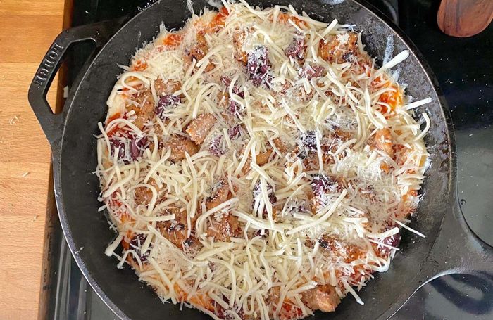 Pizza dough placed on the cheesy parmesan layer in the skillet. The pizza is topped with sausage chunks, sliced kalamata olives, shredded parmesan, and shredded mozzarella.