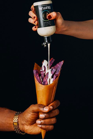 A person holding a cone of food while another person adds sauce on top.