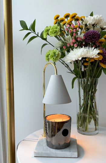 A reviewer’s candle warmer lamp sitting on a side table next to flowers and a standing lamp.