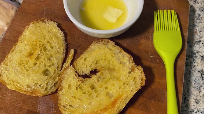 Two croissant halves placed on a cutting board, with a dish of melted butter and a pastry brush beside them.