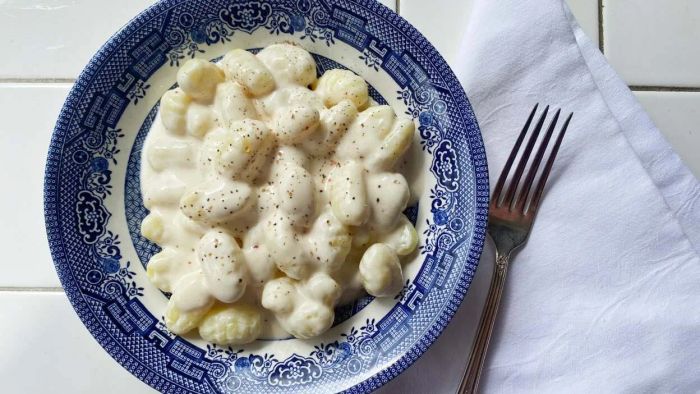 A bowl of gnocchi sits on a table in a blue china dish, covered with a creamy white sauce, speckled with black pepper.
