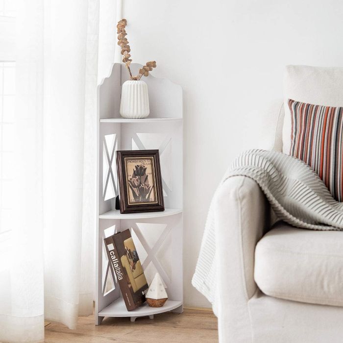 A white corner shelf unit with three levels, holding photos, books, and a faux plant, positioned next to a couch.