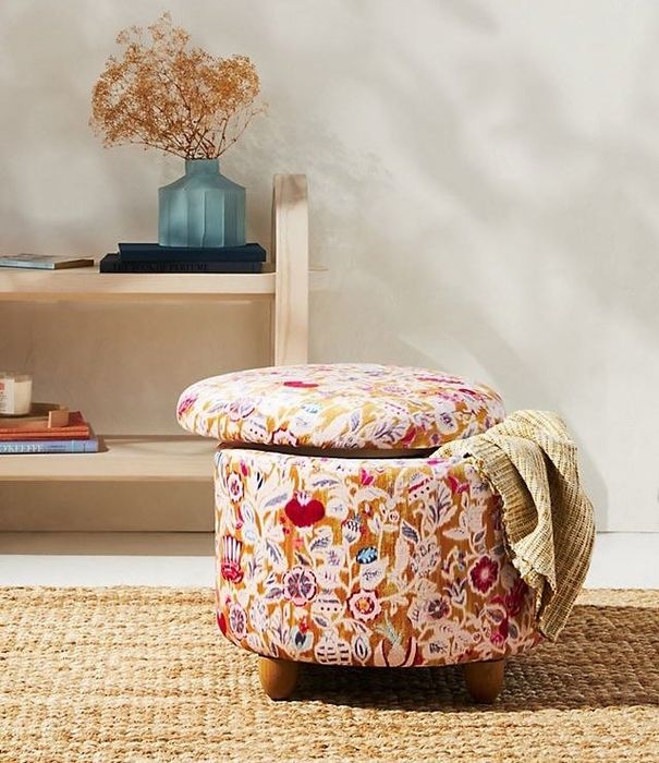 A floral-patterned storage ottoman with its lid slightly raised, and a cozy throw blanket peeking out from the inside.
