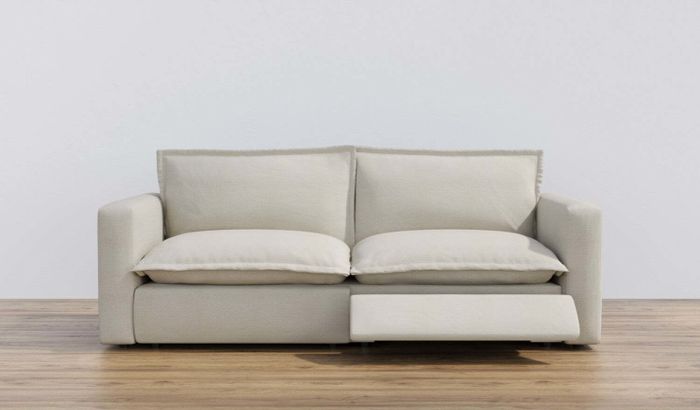 A modern beige sofa featuring two large cushions and a pull-out footrest, set against a minimalist wooden floor and a simple background.