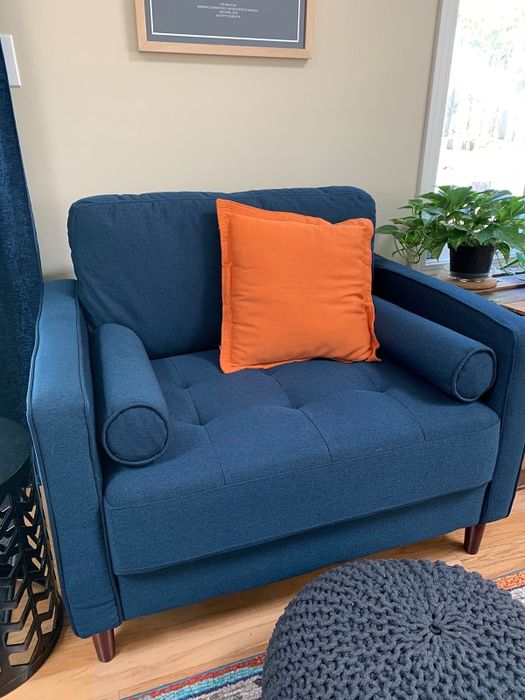 A navy blue armchair accented with a single orange cushion, set in a room adorned with plants and wall art