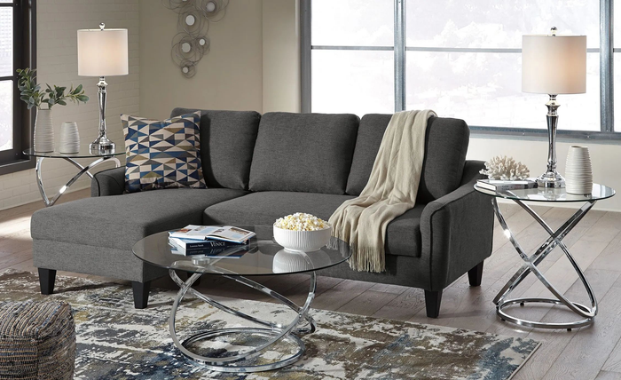 A stylish modern living room complete with a sectional sofa, geometric-patterned cushions, a throw blanket, a glass coffee table with popcorn, and chrome side tables with lamps.