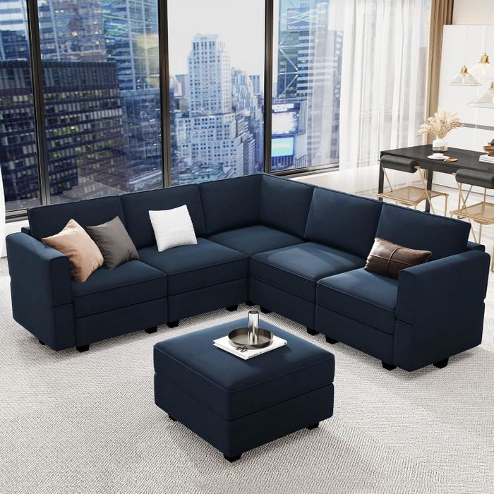 A contemporary blue sectional sofa with plush cushions and a matching ottoman is placed in a sleek urban living space with expansive windows offering a view of the city skyline