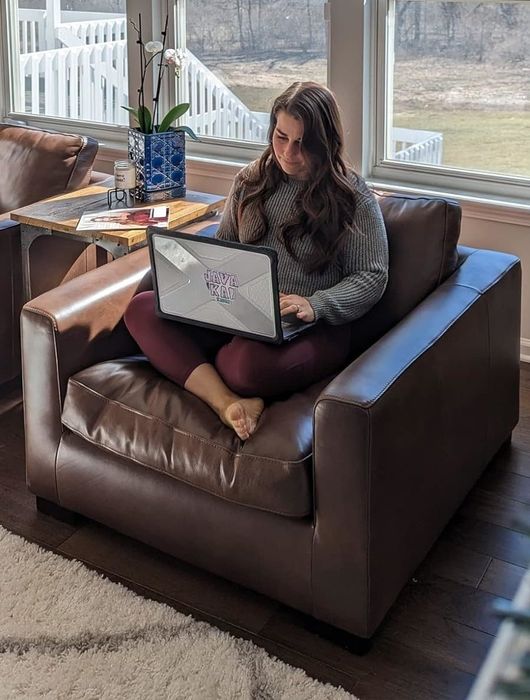 A person seated cross-legged on a brown chair, working on a laptop with a book and vase nearby on the table
