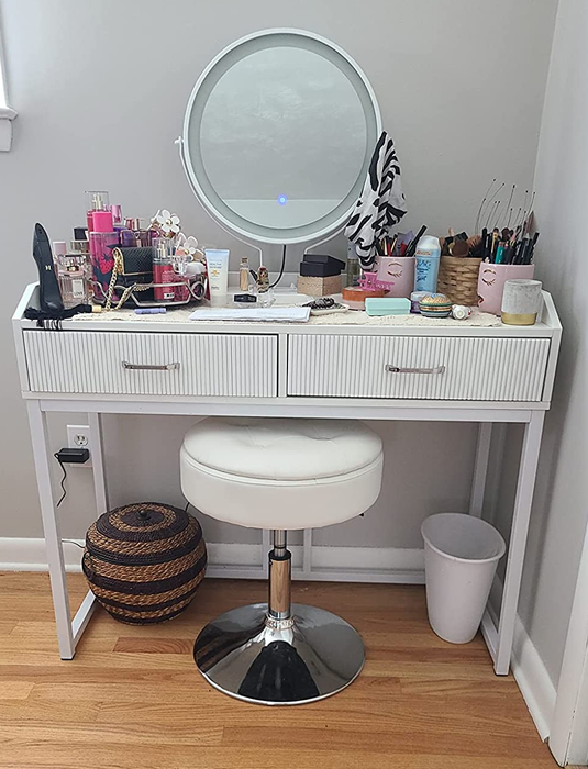 A reviewer shows an image of a white faux leather stool featuring a tufted seat, resting on a silver circular stand beneath a white vanity desk adorned with makeup.