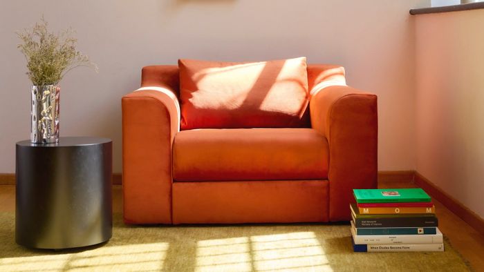 An orange armchair placed beside a round black side table holding a vase of dried flowers, with a stack of books on the floor to its right