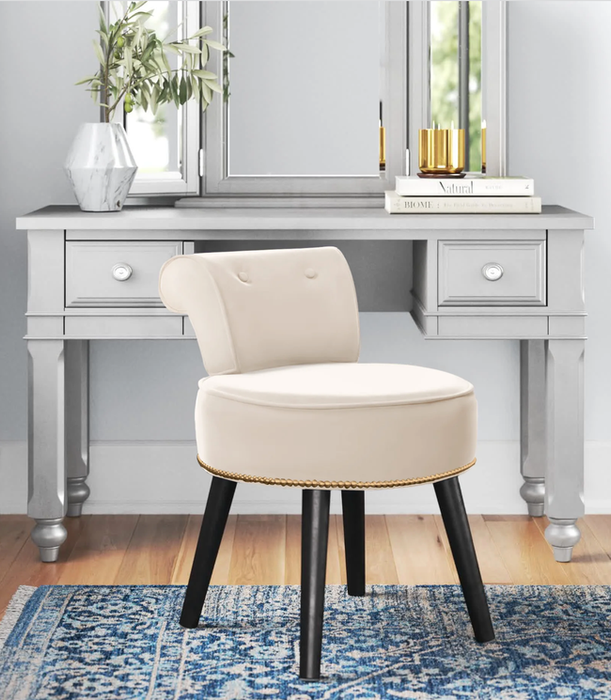 White faux leather velvet stool with black wooden legs and golden studs along the seat’s edge, placed in front of a gray vanity.