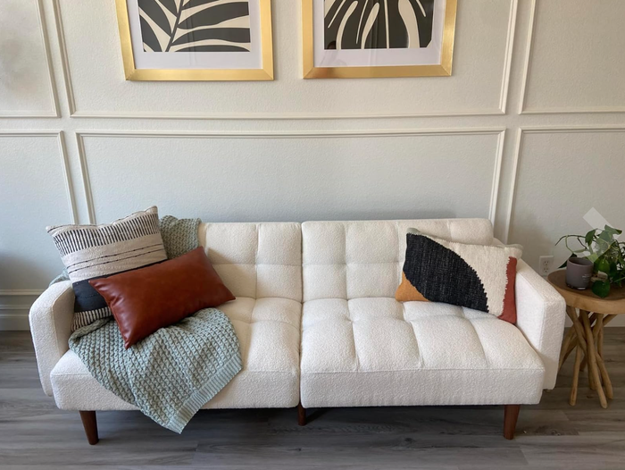 A white tufted sofa complemented by patterned pillows, a cozy blanket, a wooden side table, and framed botanical art on the wall behind.
