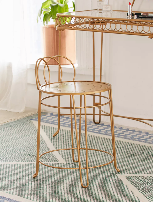 A gold short vanity stool with a looped wire backrest and legs, placed on a geometric carpet in front of a matching desk.