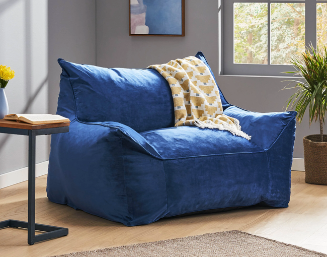 A large cobalt blue plush bean bag chair with armrests, placed on a hardwood floor with a yellow throw blanket