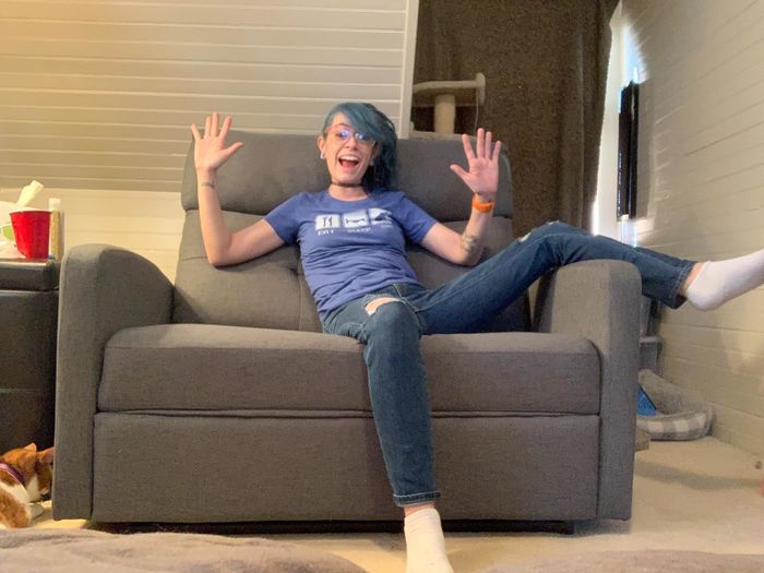 A person with blue hair sits excitedly on a gray armchair, dressed in a T-shirt and jeans. A red cup is placed on the side table, and a dog is partially visible