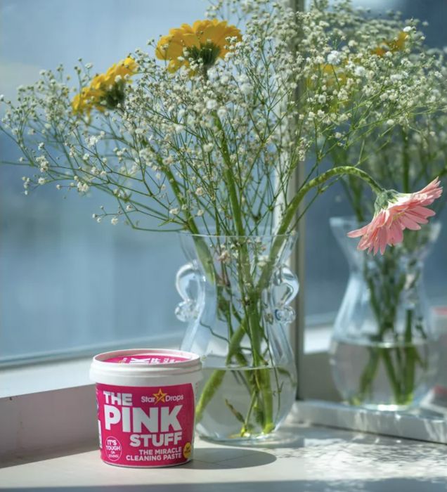 A jar of The Pink Stuff sits on a windowsill, surrounded by blooming flowers.