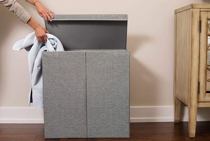 A picture of a grey laundry hamper with the lid open, as a hand places a towel into one side.