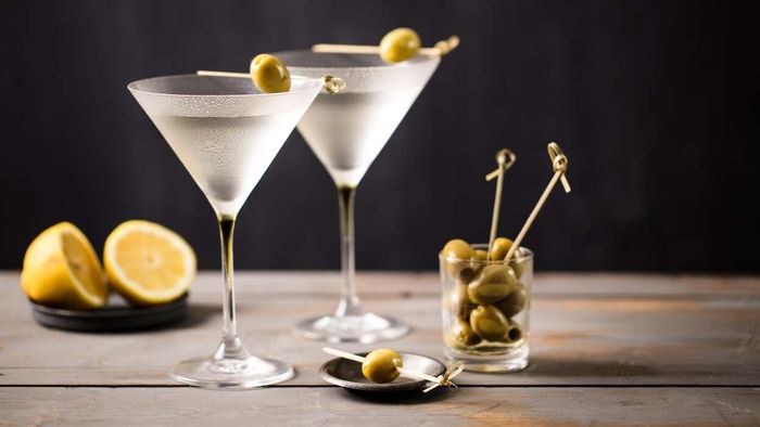 Two chilled martinis in classic martini glasses rest on a rustic wooden table, accompanied by olives and a sliced lemon.