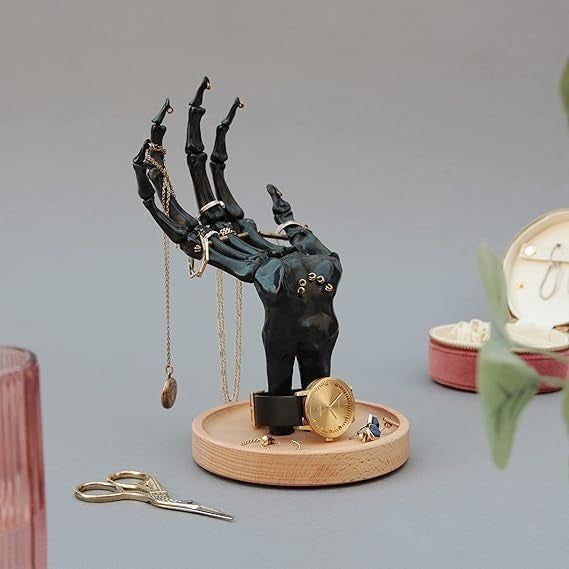 A skeleton mannequin hand jewelry stand showcasing rings, necklaces, and a watch on a wooden base. Nearby, a pair of scissors and an open jewelry box filled with various pieces