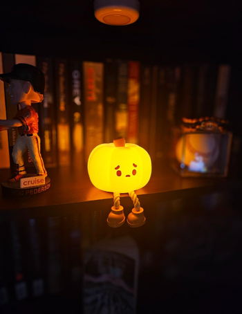 A small pumpkin-shaped night light with a face sits on a shelf surrounded by books, figurines, and a baseball.