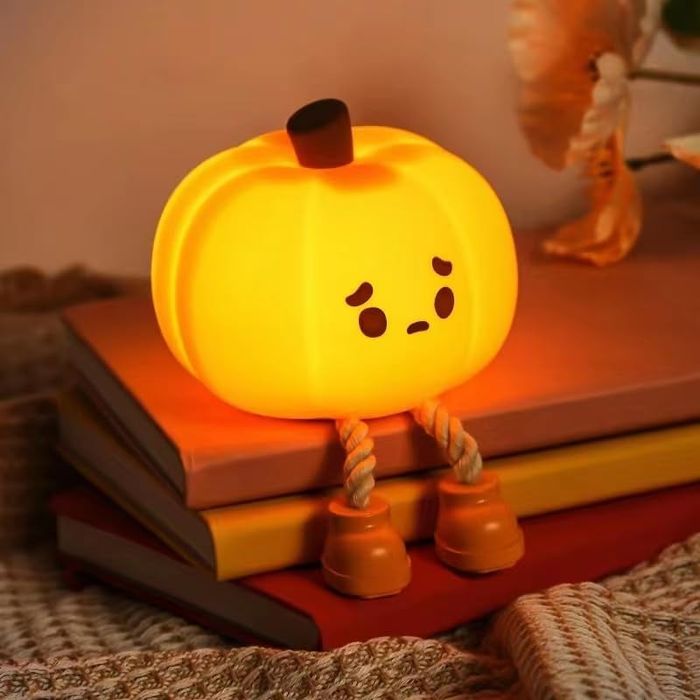 A cute pumpkin-shaped night light with a sad face, sitting atop a stack of books, complete with rope legs and shoes. A cozy and whimsical decor piece perfect for autumn.