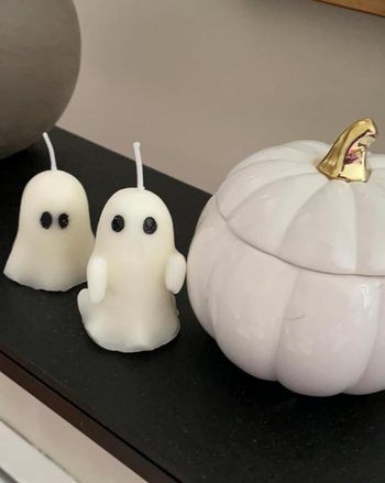 Two ghost-shaped candles with black eyes placed beside a white ceramic pumpkin featuring a golden stem, all on a black surface