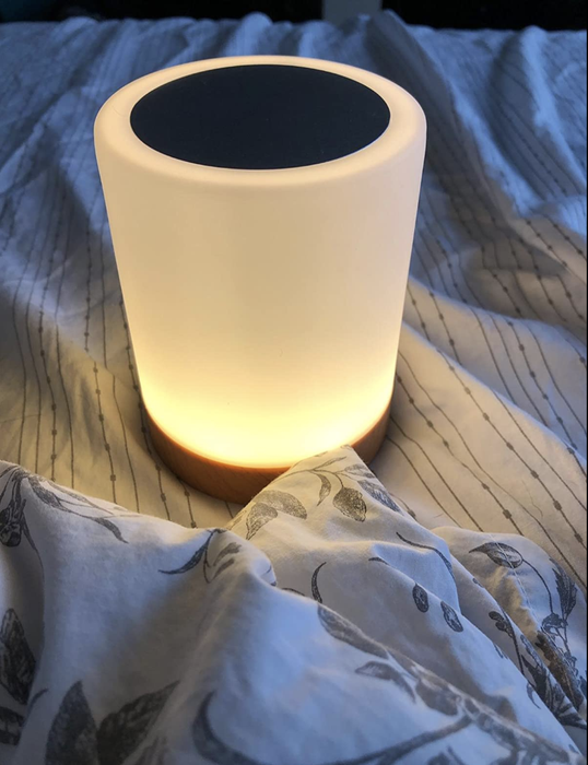 A reviewer's image of a small cylindrical lamp glowing white, placed on a bed.
