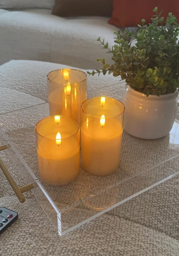 Three flickering candles and a small plant arranged on a clear tray, resting on a cushioned surface, with a remote control visible on the left