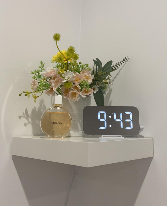 Another reviewer’s mirror alarm clock positioned on a wall-mounted corner shelf beside fresh flowers