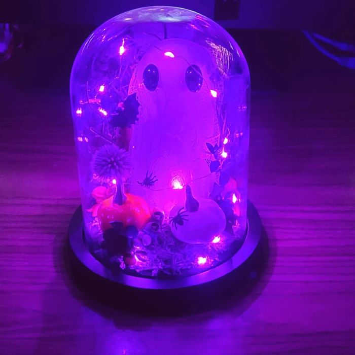 A miniature ghost figurine under a glass dome, glowing with purple lights, accompanied by small pumpkins and Halloween decor on a wooden surface