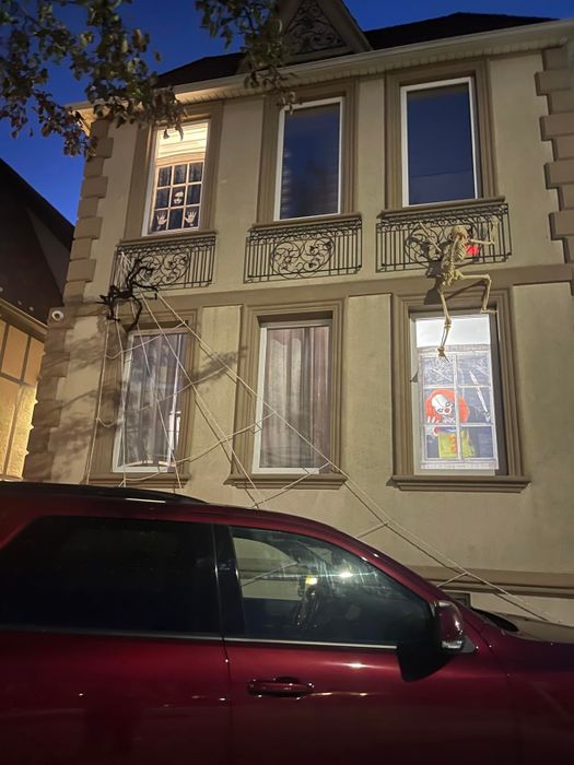 A Halloween-decorated house featuring a skeleton climbing a rope ladder to a second-story window, with spooky decorations visible in the windows