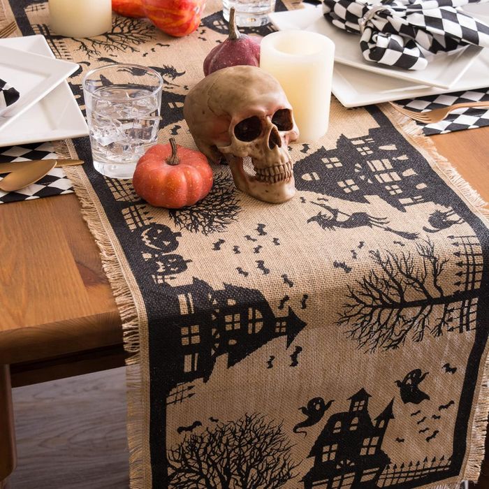 A Halloween table setup with a burlap runner featuring black spooky patterns, a skull centerpiece, candles, mini pumpkins, and festive napkins