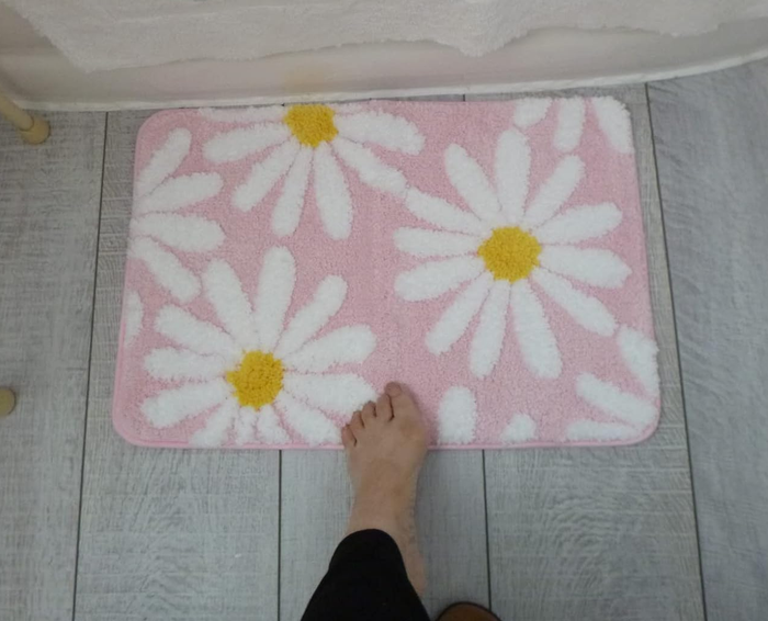 A reviewer's foot resting on the pink daisy bath mat, showcasing its soft texture and vibrant color.