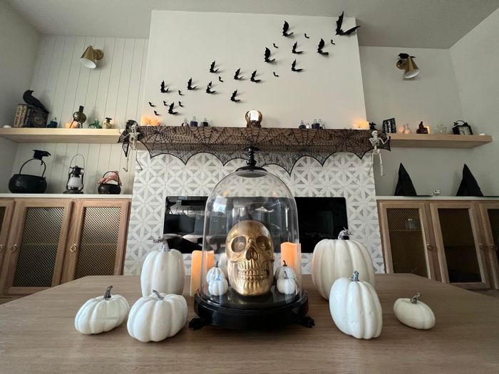 A Halloween-themed living room with a gold skull under a glass dome, surrounded by white pumpkins and candles on a table, and black bats on the wall