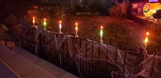 A Halloween-themed yard featuring spider webs, glowing candles on a black fence, and a lit pumpkin decoration in the background