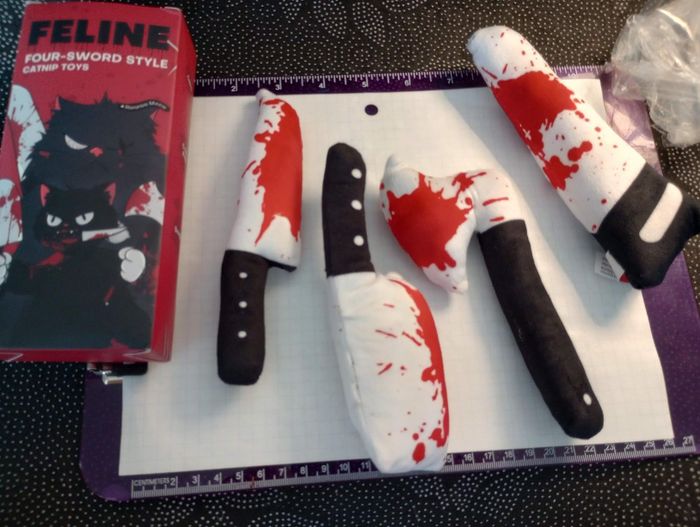 A set of catnip toys shaped like knives, featuring four plush pieces scattered on a surface, with a cat-themed packaging box in the background